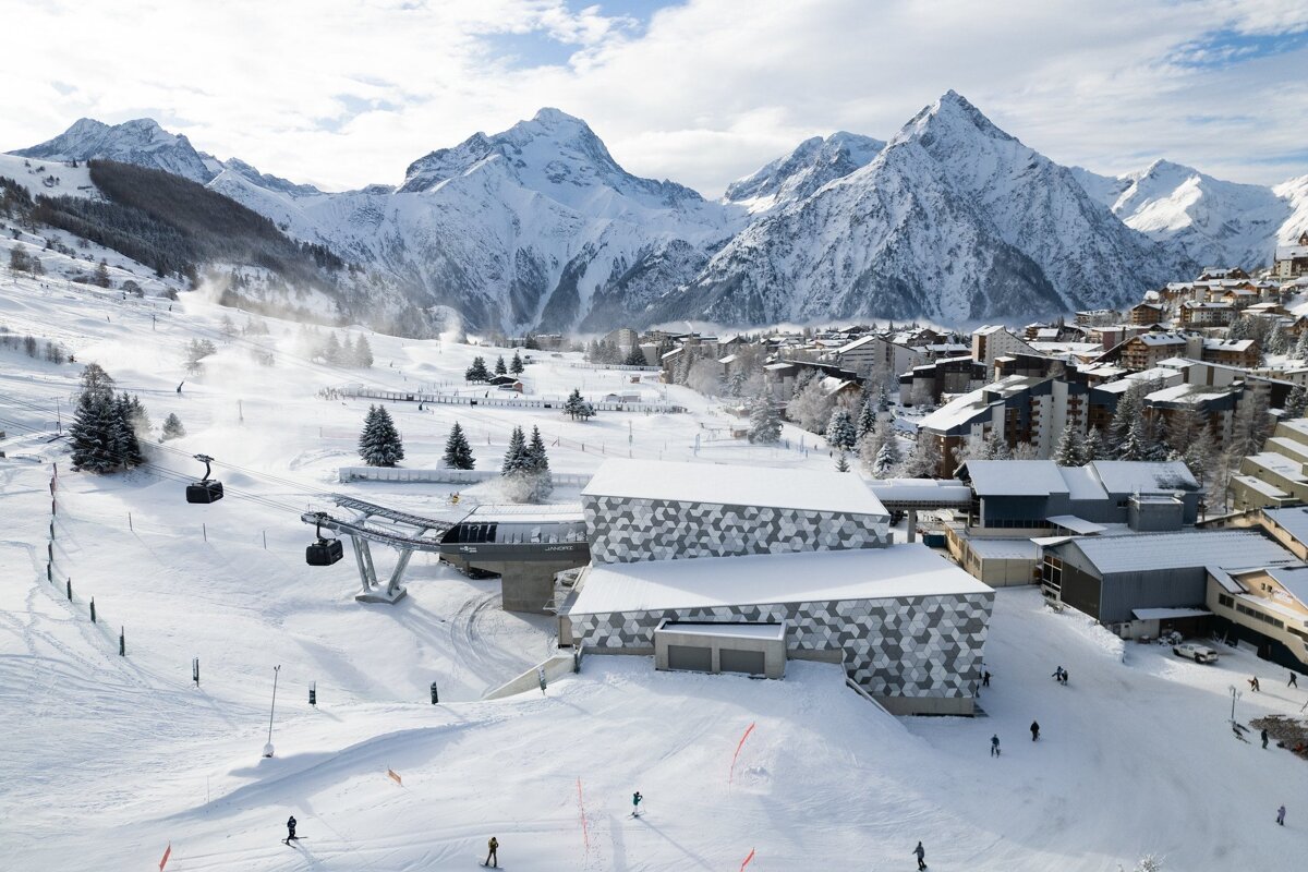 A snowy mountain ski resort with modern buildings, active ski slopes, gondola lifts, and skiers, all set against a backdrop of majestic peaks.