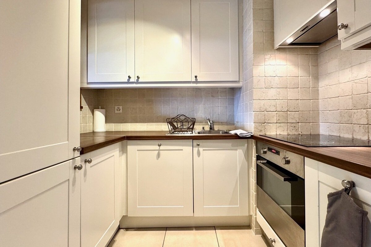 A compact, modern kitchen with white cabinets, dark wood countertops, tiled backsplash, built-in oven, hob, and sink.