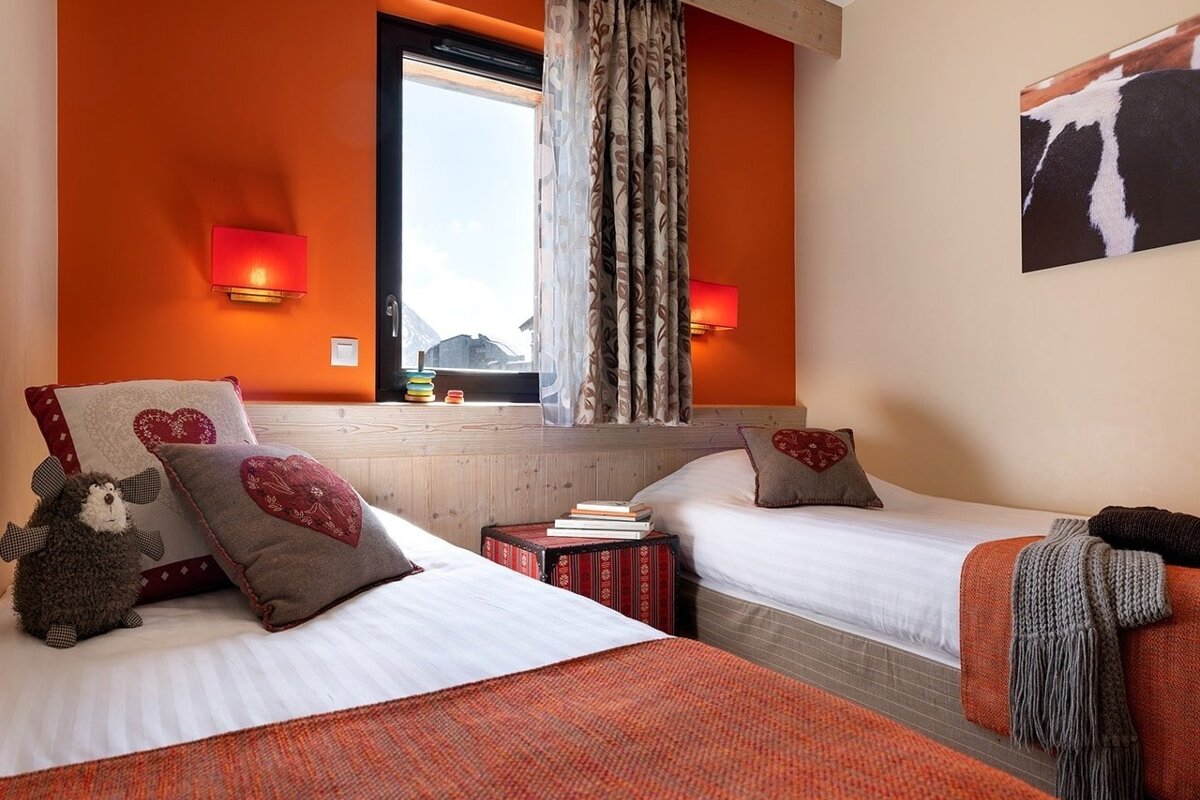 Cozy twin bedroom with bright orange walls, heart-themed pillows, a plush sheep toy, and a window view.