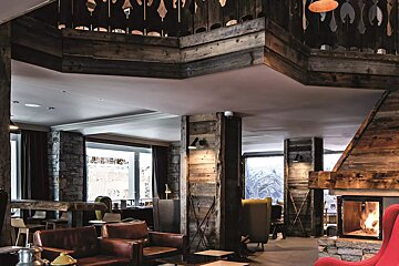 A cozy, rustic lodge interior featuring wood and stone walls, a crackling fireplace, comfortable seating, and large windows revealing an outdoor view.