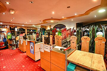 A well-lit ski and snowboard rental shop. Rows of skis, snowboards, helmets, and gear line the walls. Wooden service counters sit on a red patterned carpet.