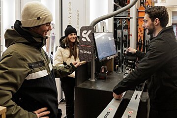 A couple in winter jackets stands at a ski service counter where a technician assists them with ski gear.