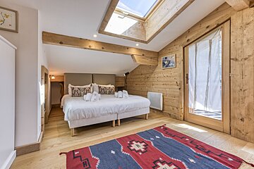 Bright, rustic bedroom featuring wood-paneled walls, exposed beams, a large bed, a skylight, and a vibrant patterned rug.
