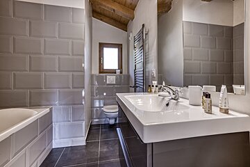 A modern bathroom features grey subway tiles, white bathtub, sink, toilet, and a small window. Wooden ceiling beams add rustic charm.