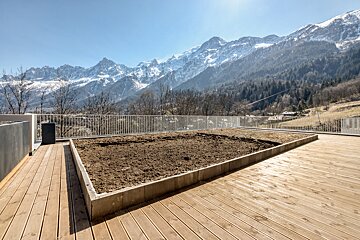 A terrace with a raised dirt bed offers a panoramic view of snow-covered mountains, a wooded valley, and a clear blue sky.