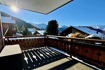 Sunny balcony view overlooking a snow-covered alpine village and majestic mountains, with bright sunlight and a clear blue sky.