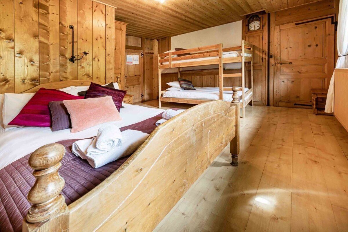 A rustic wooden bedroom features a large bed with colorful pillows in the foreground and bunk beds in the background, creating a cozy chalet atmosphere.