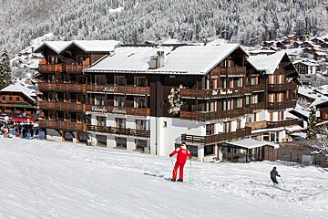 Skiers glide down a snowy slope in front of the Hotel Champs Fleuris, a multi-story alpine building with snow-covered roofs, nestled in a winter mountain landscape.