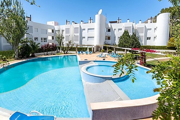 A large resort swimming pool with an attached hot tub, surrounded by white multi-story buildings and green plants under a clear blue sky.