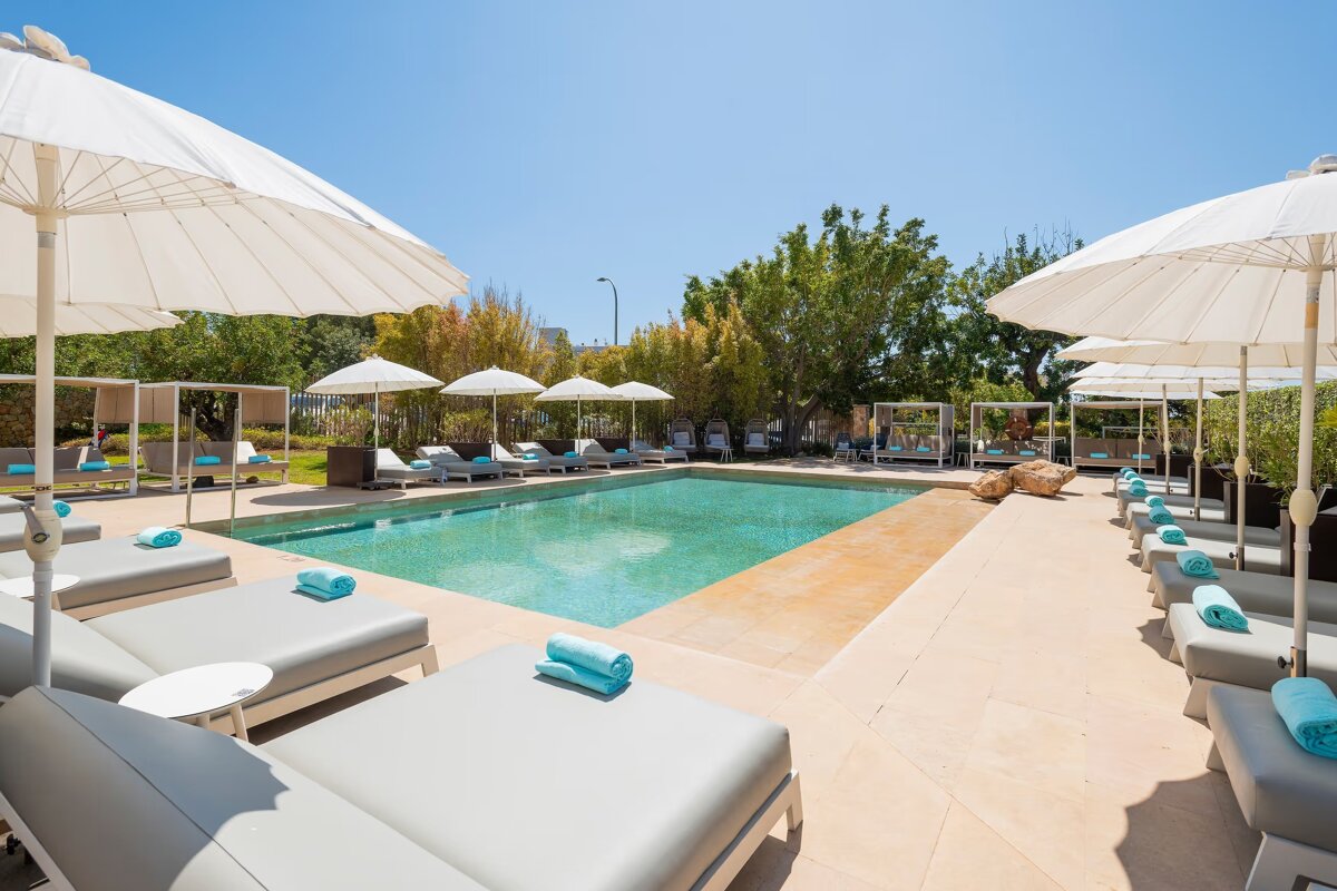 A serene resort pool area with numerous grey lounge chairs, turquoise towels, and white umbrellas under a bright, clear sky, surrounded by greenery.