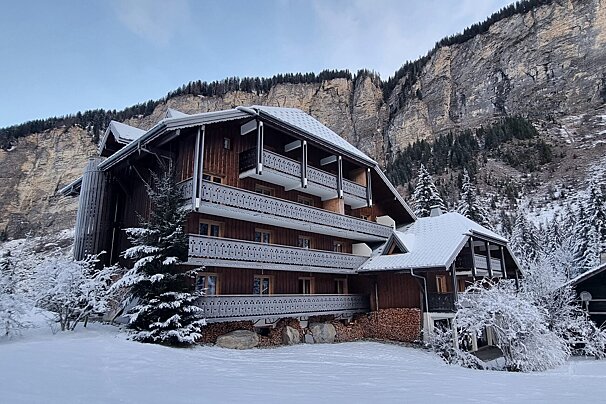 A large, snowy wooden chalet stands in a winter wonderland, backed by towering cliffs and snow-covered pine trees.