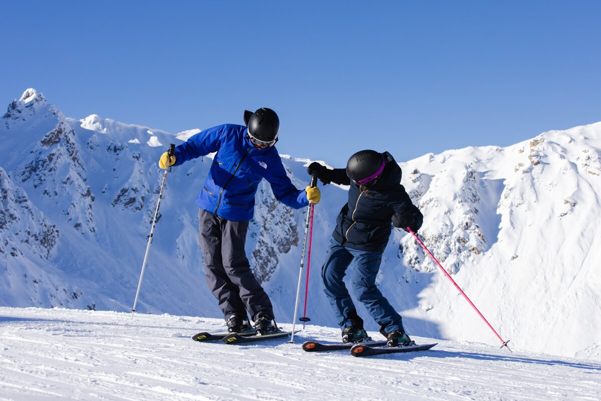 A man in a blue jacket helps a woman ski down a snowy slope