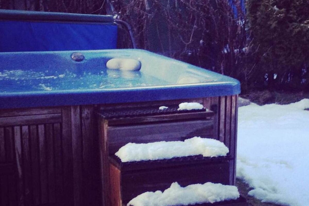 An outdoor blue hot tub with wooden siding, partially covered in snow. Its steps and the ground around it are also snowy.