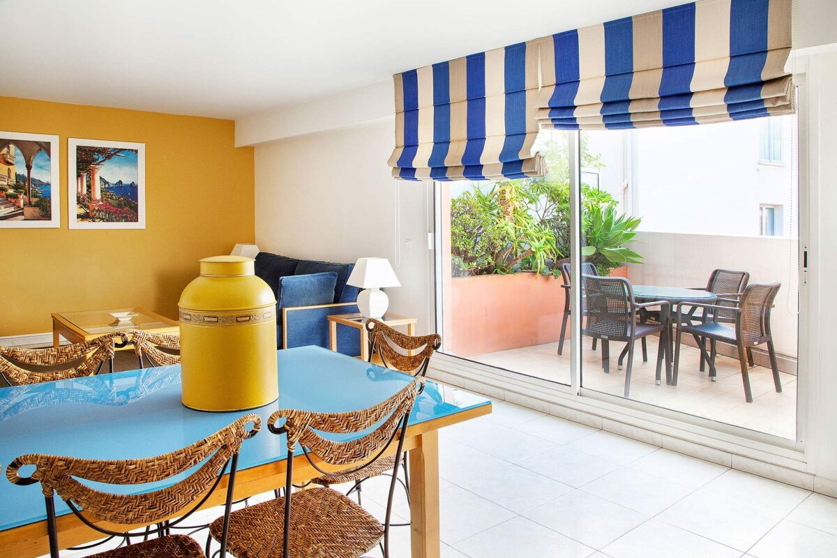 A blue table with wicker chairs and a yellow vase on it