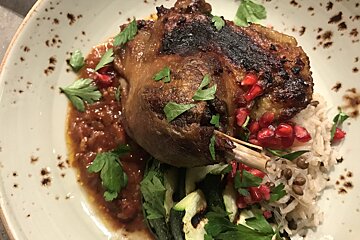 A crispy-skinned duck leg with vibrant pomegranate seeds, fresh parsley, and a savory sauce, served with rice, lentils, and grilled zucchini.
