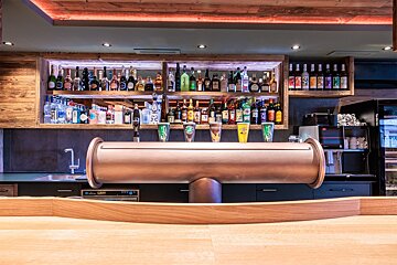 A modern, well-stocked bar features numerous liquor bottles on wooden shelves, a prominent copper beer tap, and glasses, illuminated by warm lights.