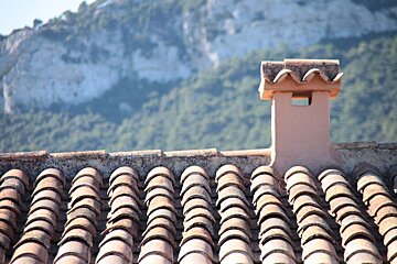A rustic tiled roof with an orange chimney stands out against a blurred backdrop of green, rocky mountains under a bright sky.