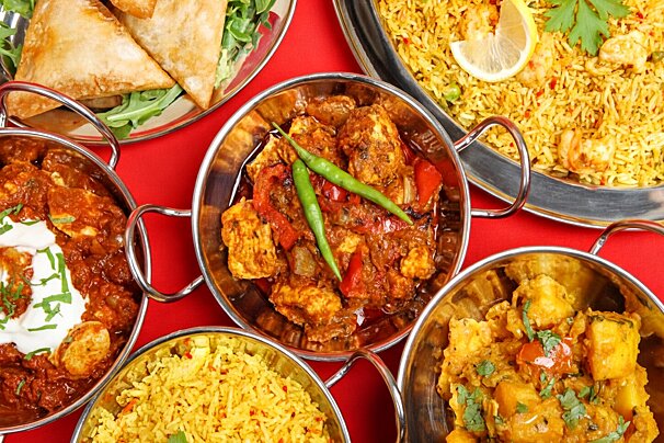 A vibrant spread of Indian cuisine on a red tablecloth, featuring various curries in metal bowls, samosas, and shrimp biryani with lemon.