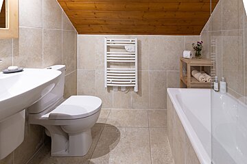 A modern bathroom with beige tiled walls and floor, a sloped wooden ceiling, sink, toilet, bathtub, and a white heated towel rail.