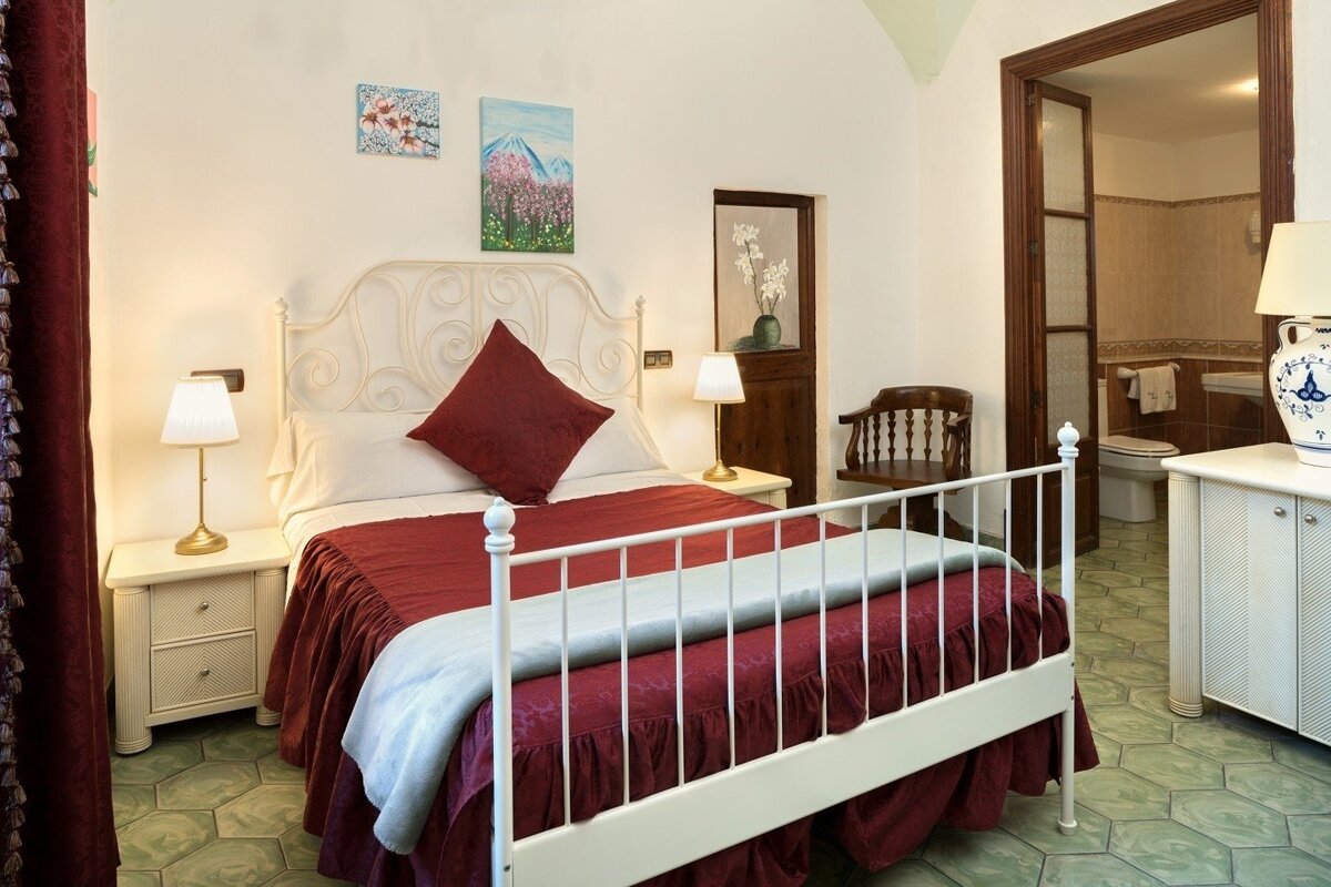 A cozy bedroom with a white wrought iron bed, red bedding, and unique green hexagonal floor tiles. An open doorway reveals an adjacent bathroom.