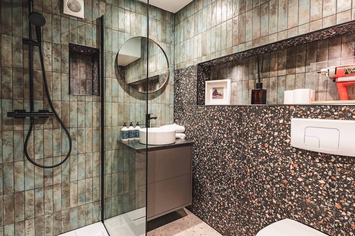 A modern bathroom with a shower, sink, and toilet. Distinctive green/brown vertical tiles and speckled terrazzo create a textured, contemporary look.