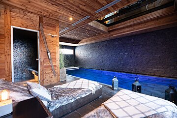 A luxurious chalet-style indoor pool with wooden walls/ceiling, stone accent wall, bright blue water, and cozy lounge seating with fur throws.