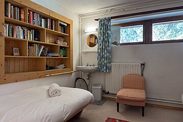 A compact room with a single bed, built-in wooden bookshelf, sink vanity with a mirror, and high windows overlooking trees. An armchair sits beside a radiator.