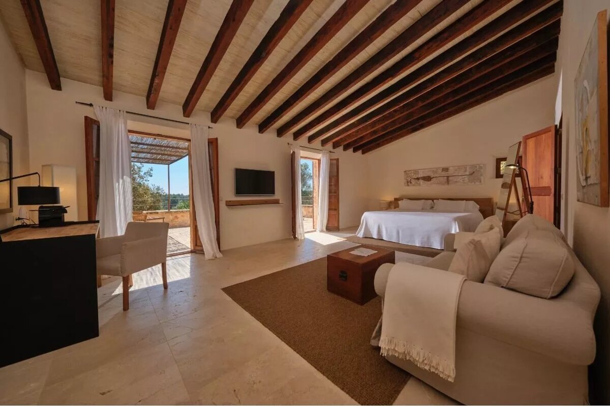 Bright, rustic bedroom with wood beams, large bed, seating area, TV, and open doors to a sunny terrace.