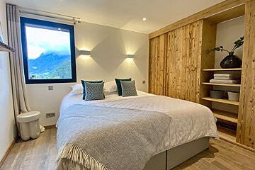 A cozy bedroom with a large bed, teal cushions, a big window revealing mountain views, and a rustic wooden wardrobe with open shelves.
