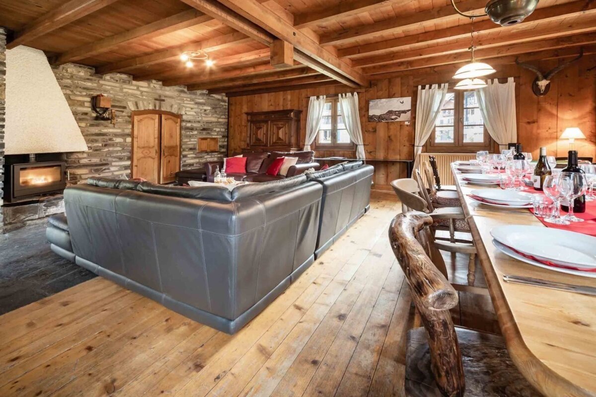 A rustic, wood-paneled chalet interior with a large stone fireplace, comfy L-shaped sofa, and a fully set dining table, offering a cozy mountain retreat.