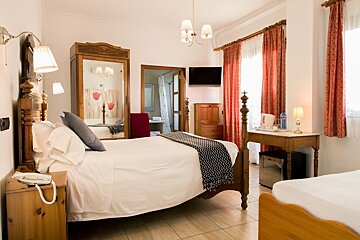 Cozy, traditionally furnished hotel bedroom with two beds (one four-poster), white linens, red patterned curtains, wooden furniture, and a bathroom.