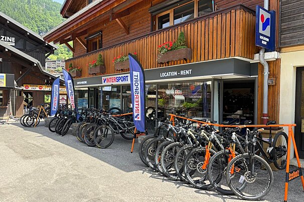 A sunny outdoor scene shows an Intersport bike rental shop with a wooden facade, featuring many mountain bikes parked outside and mountains in the background.