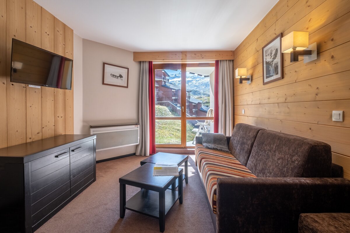Cozy chalet living room with a brown sofa, wooden paneled walls, a mounted TV, and a glass door opening to scenic mountain views.