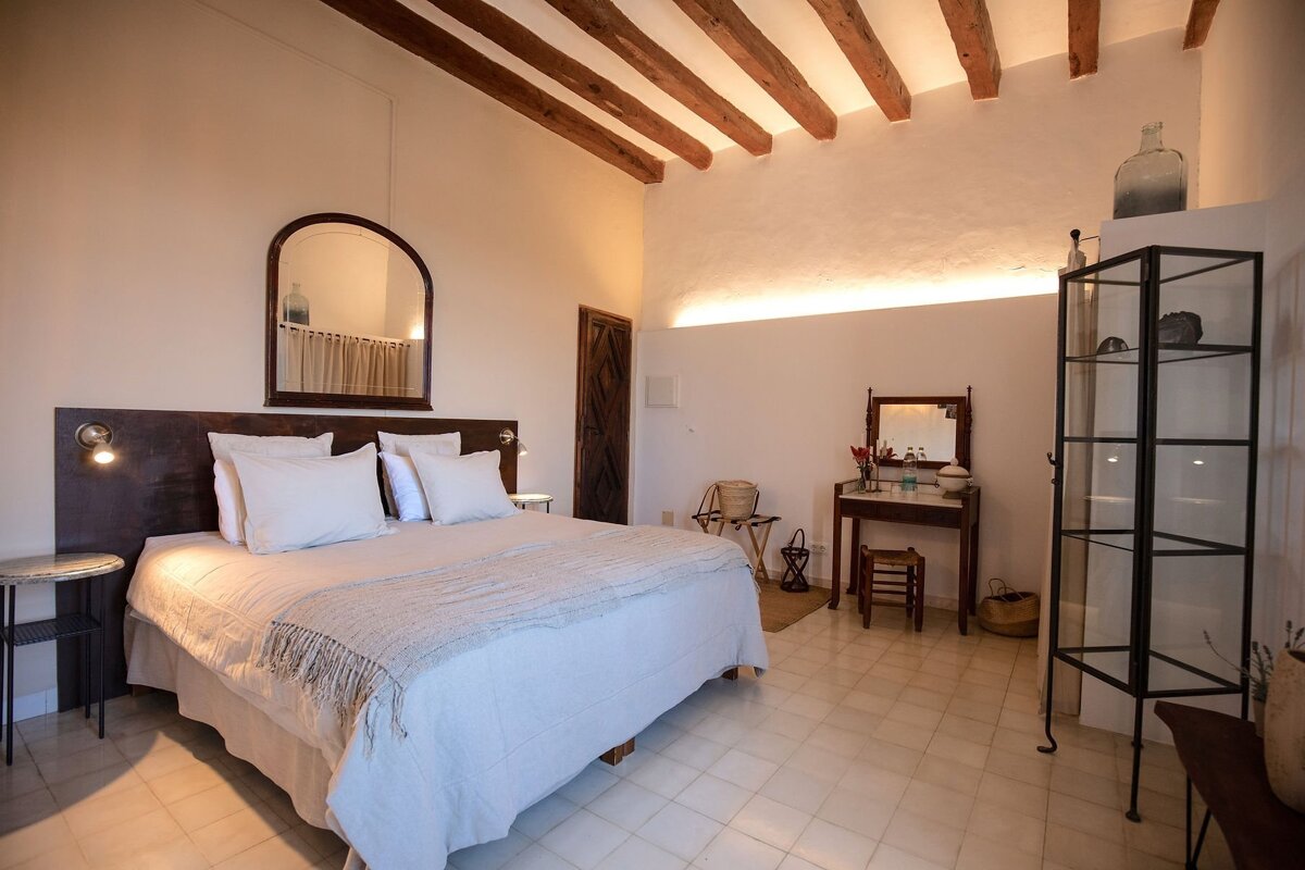 A rustic, Mediterranean-style bedroom featuring a large bed, exposed wooden beams, arched mirror, vanity, and warm cove lighting.