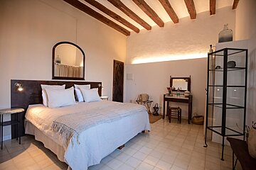 A rustic, Mediterranean-style bedroom featuring a large bed, exposed wooden beams, arched mirror, vanity, and warm cove lighting.