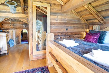 A cozy, rustic wooden bedroom with a bed, faux fur, bright pillows, and a large mirror. A wooden loft/hallway is visible.