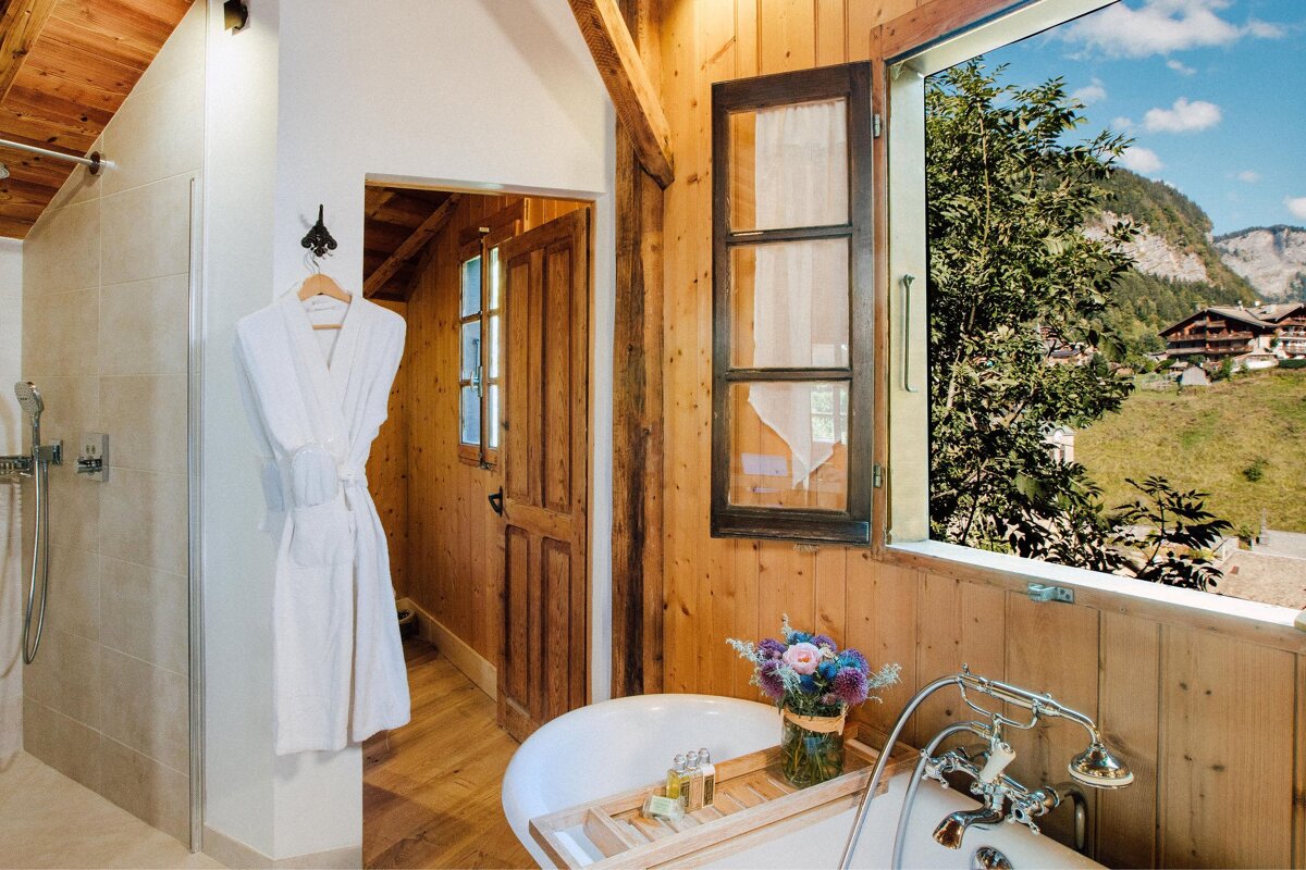Rustic bathroom with a white tub, bathrobe, and a scenic mountain village view through an open window, featuring warm wood paneling.