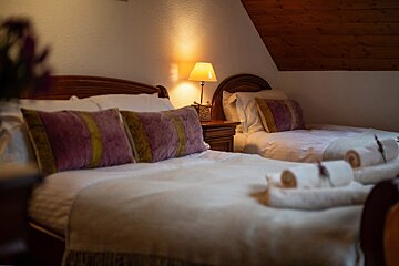 A cozy, warm-lit bedroom features two beds (one large, one small), ornate wooden furniture, purple and gold pillows, and fresh white towels. A sloped wooden ceiling adds charm.