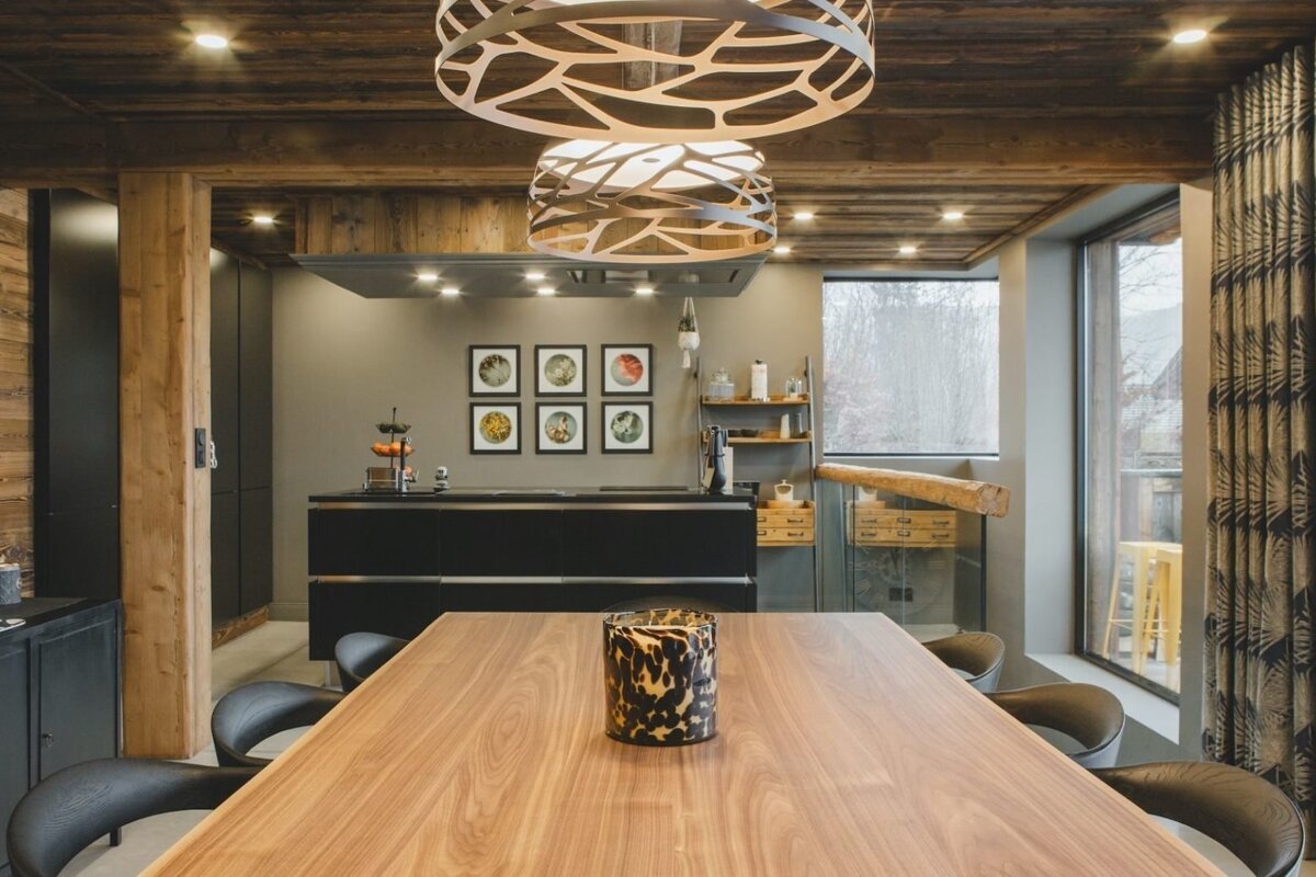 A chic dining room and kitchen with a long wood table, modern black cabinetry, rustic wooden ceiling/beams, and unique circular chandeliers. Large windows offer a view.