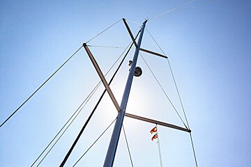 Low-angle shot looking up at a sailboat mast and rigging with small flags, set against a bright, clear blue sky.