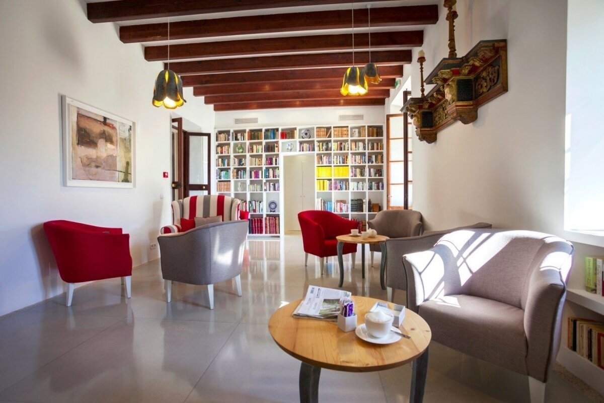 A bright, inviting library lounge with floor-to-ceiling bookshelves, comfy armchairs in red and grey, coffee tables, and rustic wooden beams.