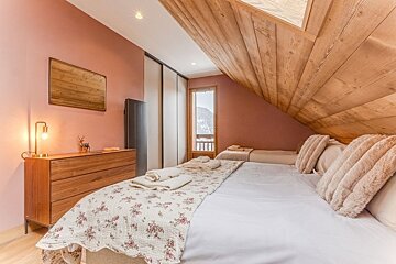 Cozy attic bedroom with slanted wood ceilings, pink walls, a large bed, a wooden dresser, and a door showing snowy mountain views.