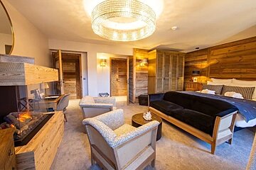 A rustic hotel suite with a cozy fireplace, two armchairs, a black sofa, a double bed, and warm wooden paneling.