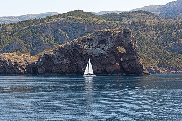 A white sailboat sails on calm blue water near a massive, arched rock formation, with forested mountains in the background.
