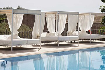 Three white canopied daybeds with sheer curtains line a serene pool, reflecting in the blue water, creating a luxurious resort scene.