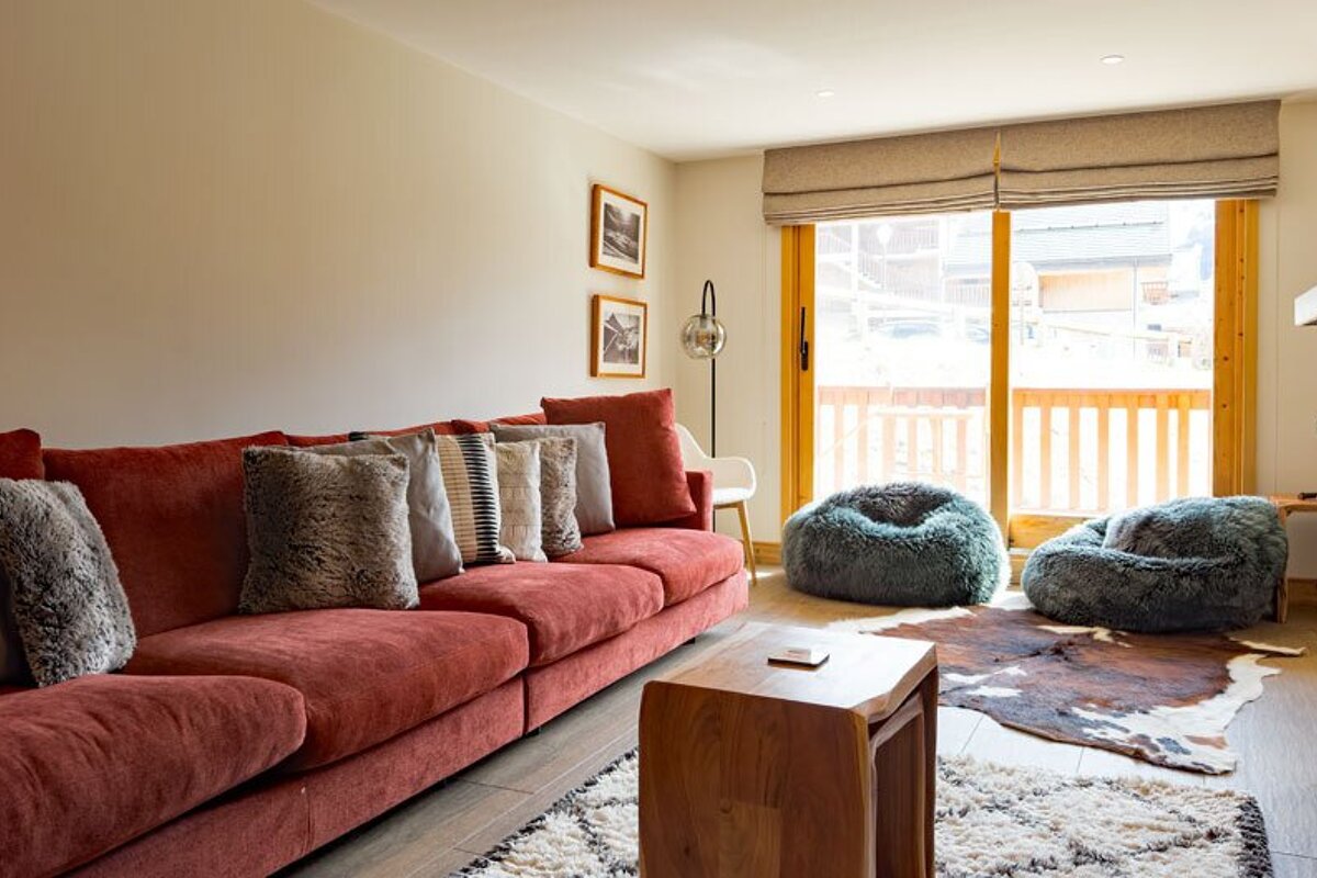 A cozy living room featuring a long red sofa, furry bean bags on a cowhide rug, and a large glass door opening to an outdoor view.