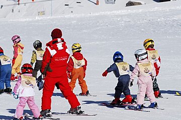 A ski instructor in a red suit leads a group of young children in a ski lesson on a sunny, snowy mountain slope at Val Thorens.