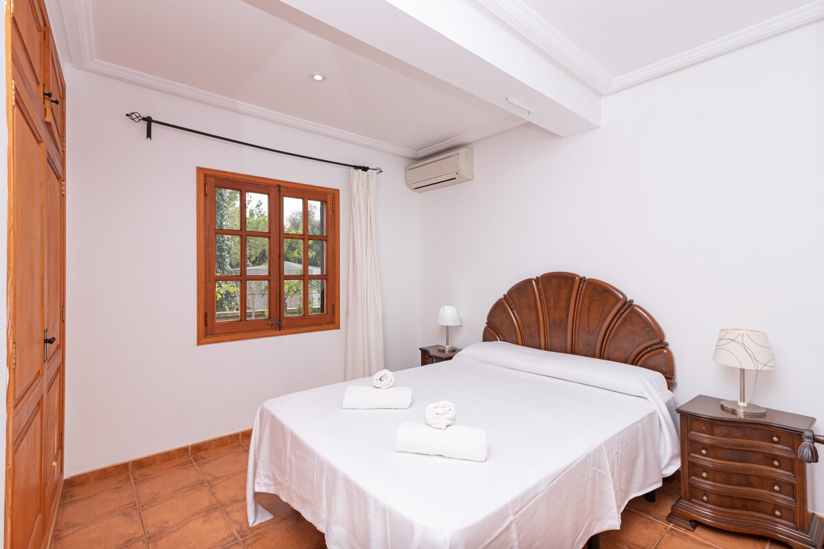 A cozy bedroom features a wooden shell headboard bed with white linen, twin nightstands with lamps, a wood-framed window, terracotta floors, and an AC unit.