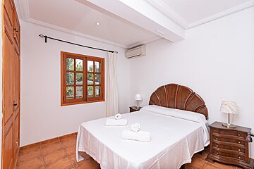 A cozy bedroom features a wooden shell headboard bed with white linen, twin nightstands with lamps, a wood-framed window, terracotta floors, and an AC unit.