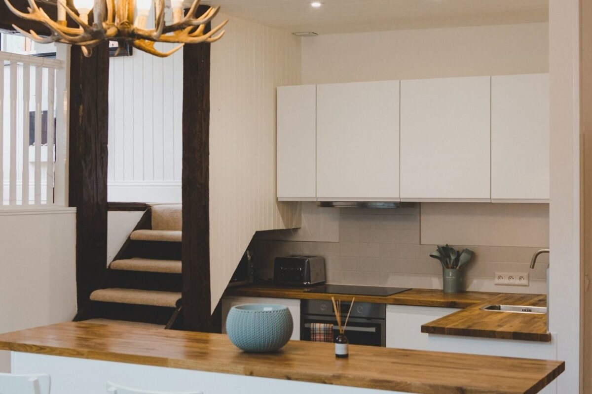 A bright, modern kitchen with white cabinets, wood countertops, and a breakfast bar. A carpeted staircase with dark wood beams and an antler chandelier are visible.